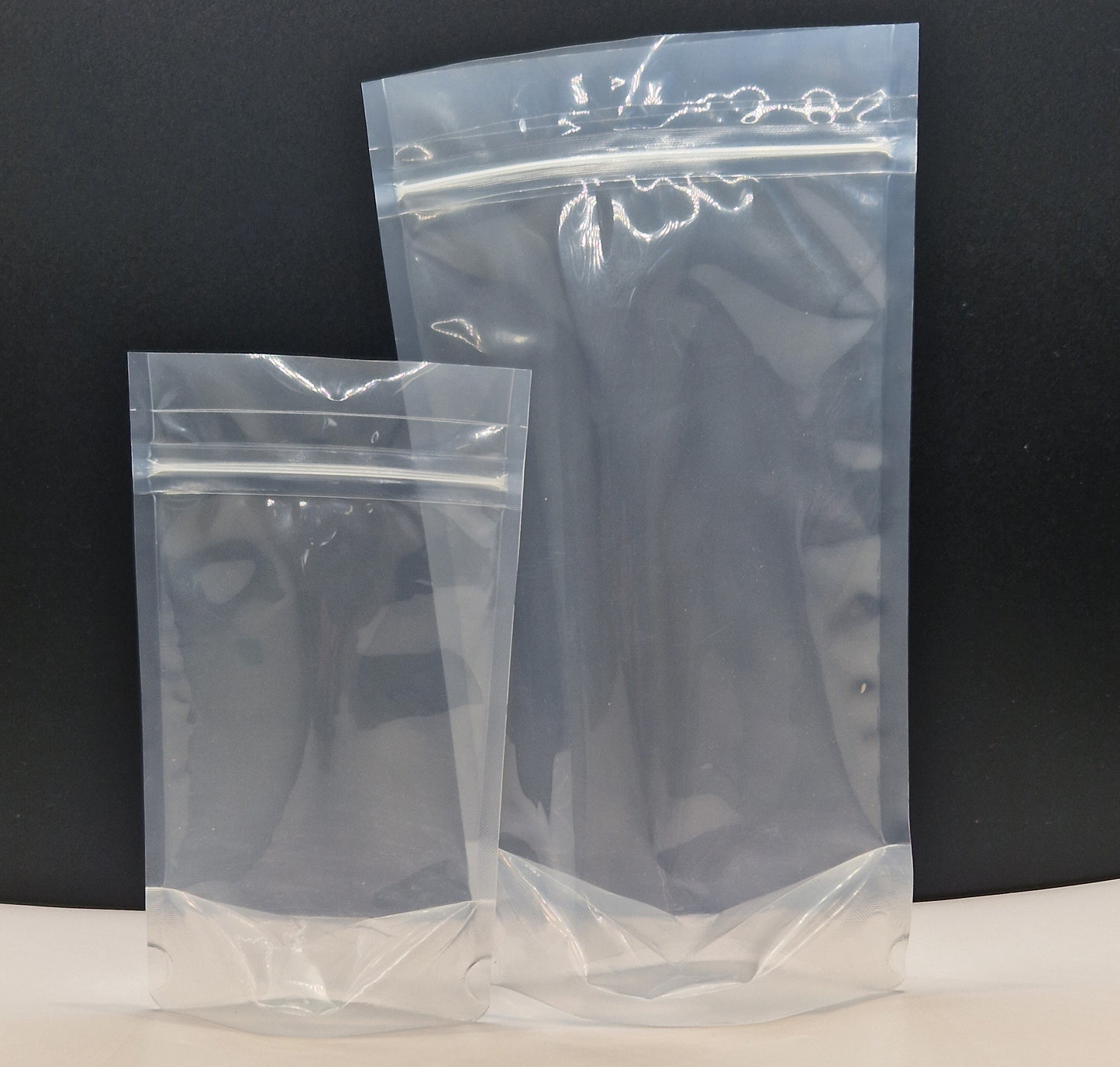 Two clear recyclable resealable stand up pouches of different sizes, displayed empty against a black background.