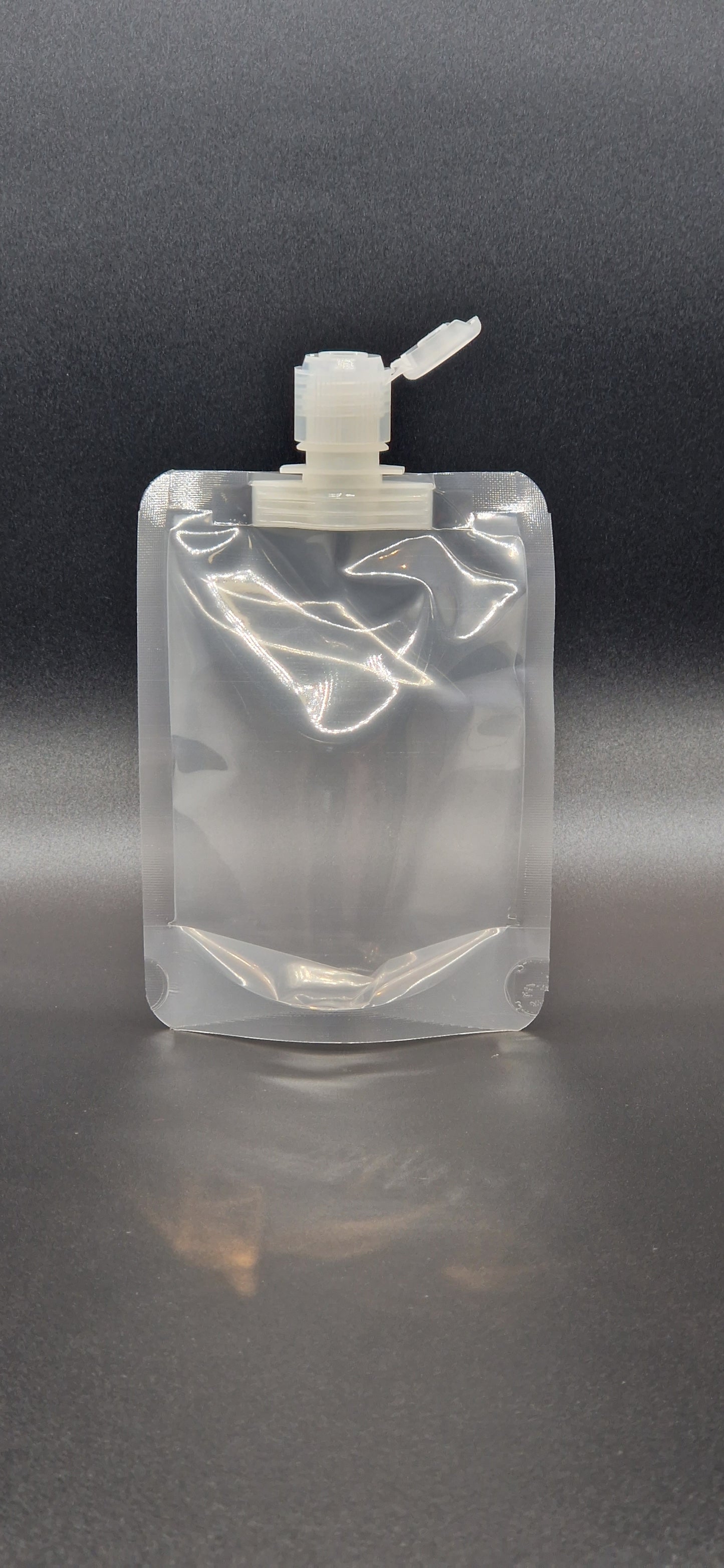 Clear plastic spout pouch with centre spout and flip-top cap, shown upright against a dark gradient background.