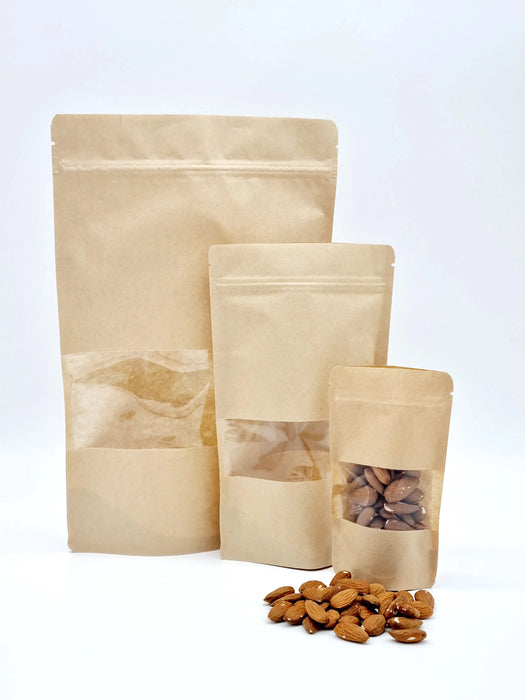 3 different sized brown kraft stand up pouches with window with a pile of uts in front of them.