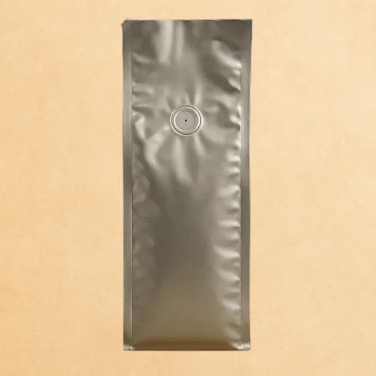 Flat bottom silver coffee bag made from foil, featuring a one-way valve and open top, shown upright against a beige background.