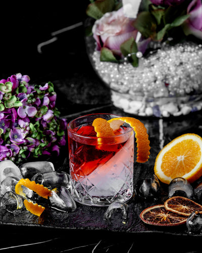 Cocktail with orange garnish on a dark surface with flowers and ice cubes.