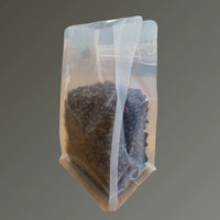 Clear recyclable flat bottom bag with Rip n Zip and open top, filled with coffee beans, shown from a side angle against a grey background.