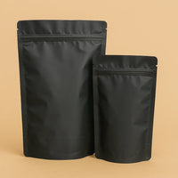 Two matt black foil stand up pouches with resealable press-to-close strips, shown upright against a beige background.